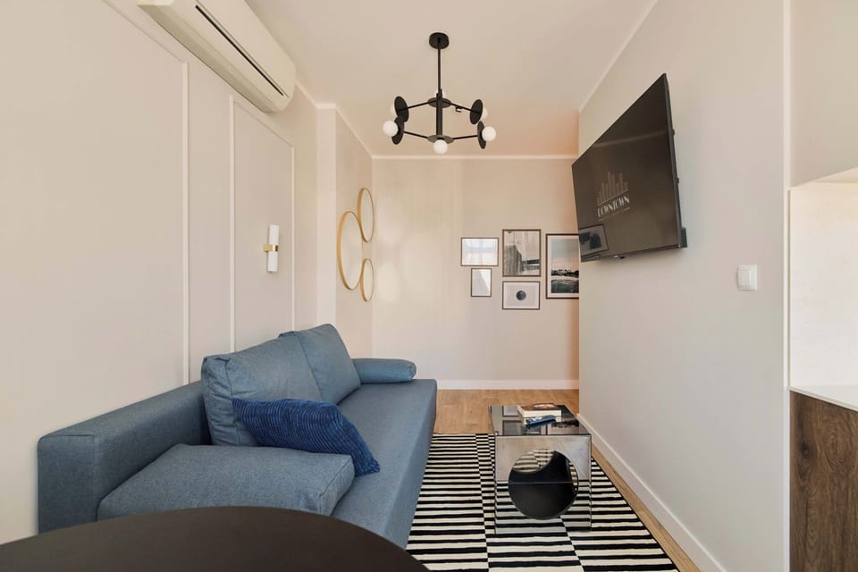 Stylowy i przytulny salon z wygodną sofą, dużym telewizorem i eleganckimi dodatkami. Jasna kolorystyka, drewniana podłoga, dekoracyjne lustra oraz nastrojowe oświetlenie tworzą komfortową przestrzeń do wypoczynku i spędzania czasu.