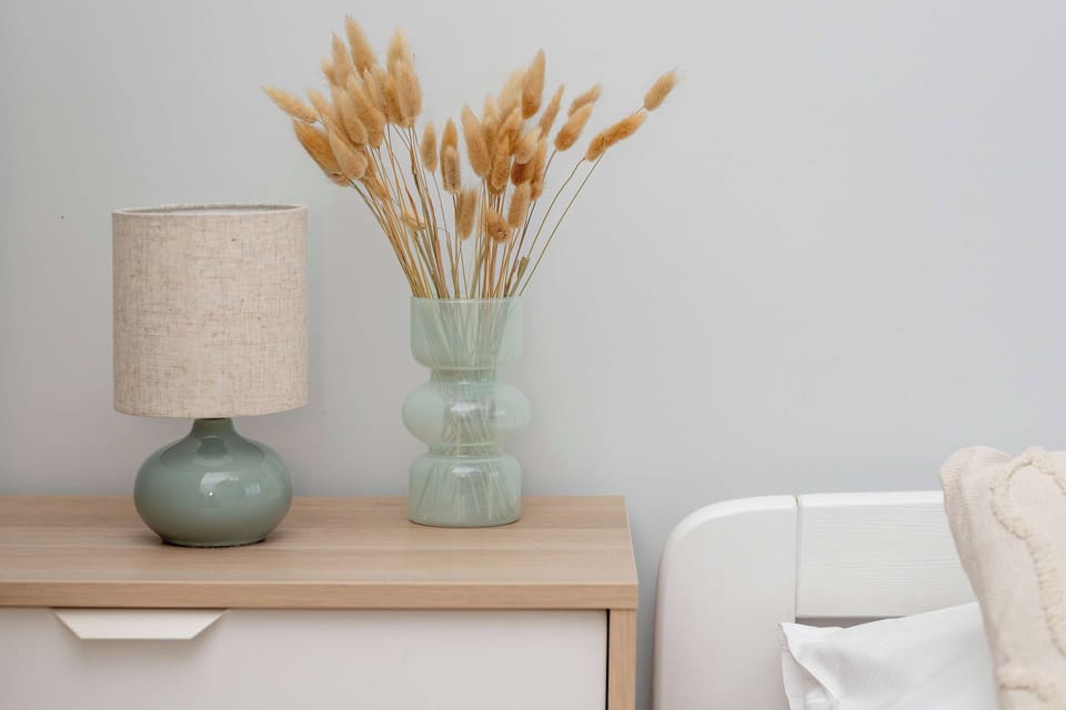 A wooden nightstand holds a petite table lamp with a dark base and light shade. Beside it stands a clear glass vase filled with dried floral stems.