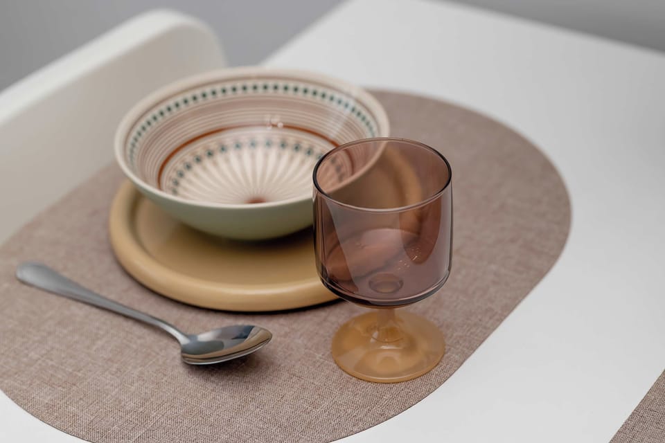A dining place setting rests neatly on an oval placemat atop a white table. It features a ceramic bowl on a wooden plate, a spoon, and a stemmed glass.