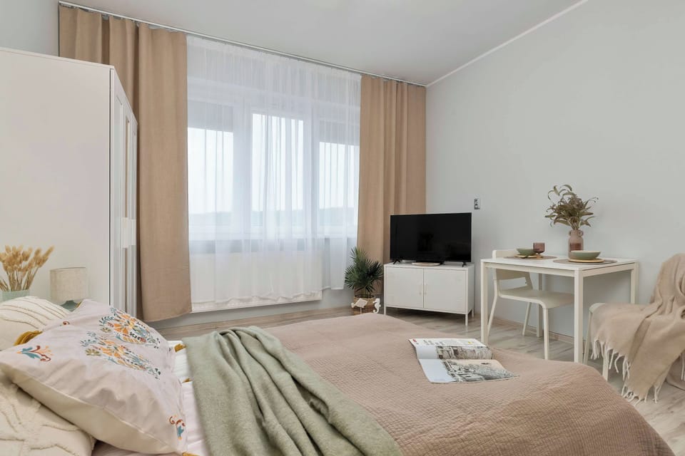 Daylight illuminates the bedroom through large windows dressed with sheer curtains. The room includes a double bed and a TV corner console.