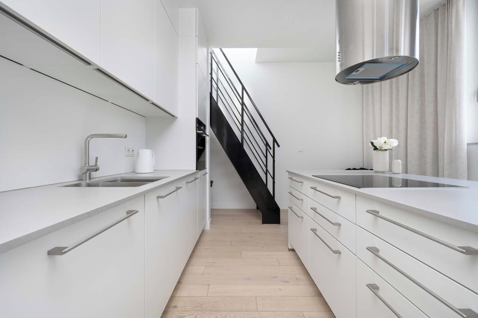 A modern kitchen is fitted with sleek white cabinetry, integrated appliances, and a minimalist black-and-wood staircase leading upstairs. Clean lines and smooth countertops emphasize the apartment's contemporary feel.