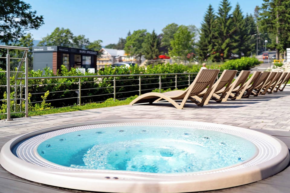 A round outdoor jacuzzi filled with bubbling water, surrounded by sun loungers on a spacious pool deck. Trees and landscaped greenery provide a peaceful setting.