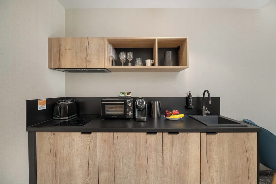 A compact kitchen with wood-effect cabinetry, a dark countertop, and open shelving. Small appliances and decorative items are arranged along the work surface.