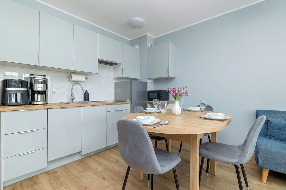 An kitchenette and dining area includes light grey cabinetry and modern appliances. A round wooden dining table surrounded by grey chairs sits in the center of the room.