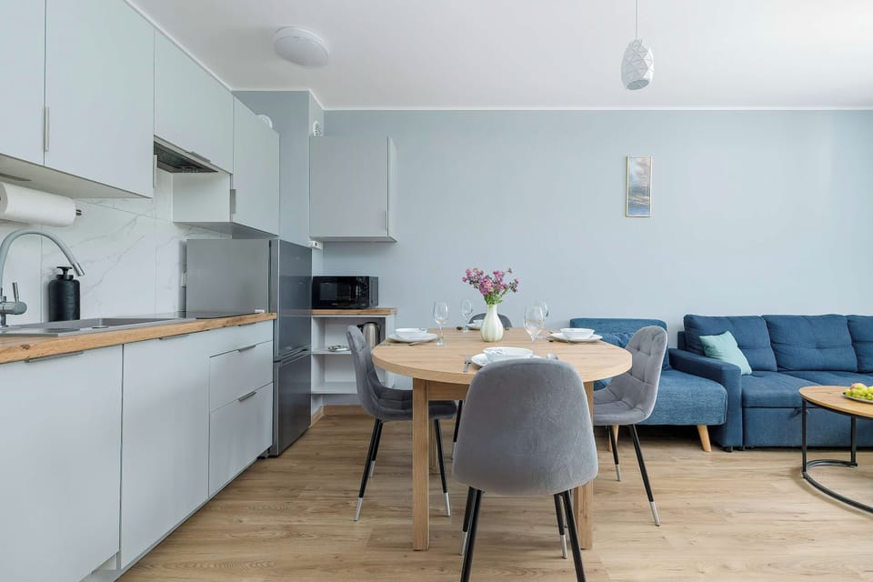 A bright kitchen and dining space features a round wooden table accompanied by grey upholstered chairs. Light grey kitchen cabinets run along the back wall under recessed ceiling lights.