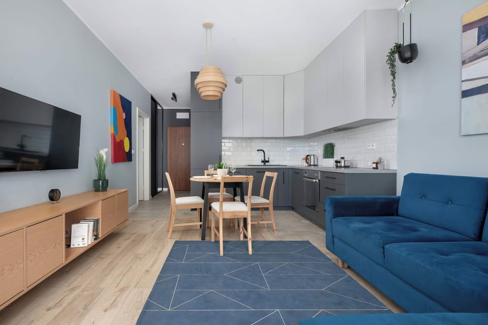A modern open-plan living area with a blue sofa, dining table, kitchen, and TV. Large windows provide plenty of natural light.