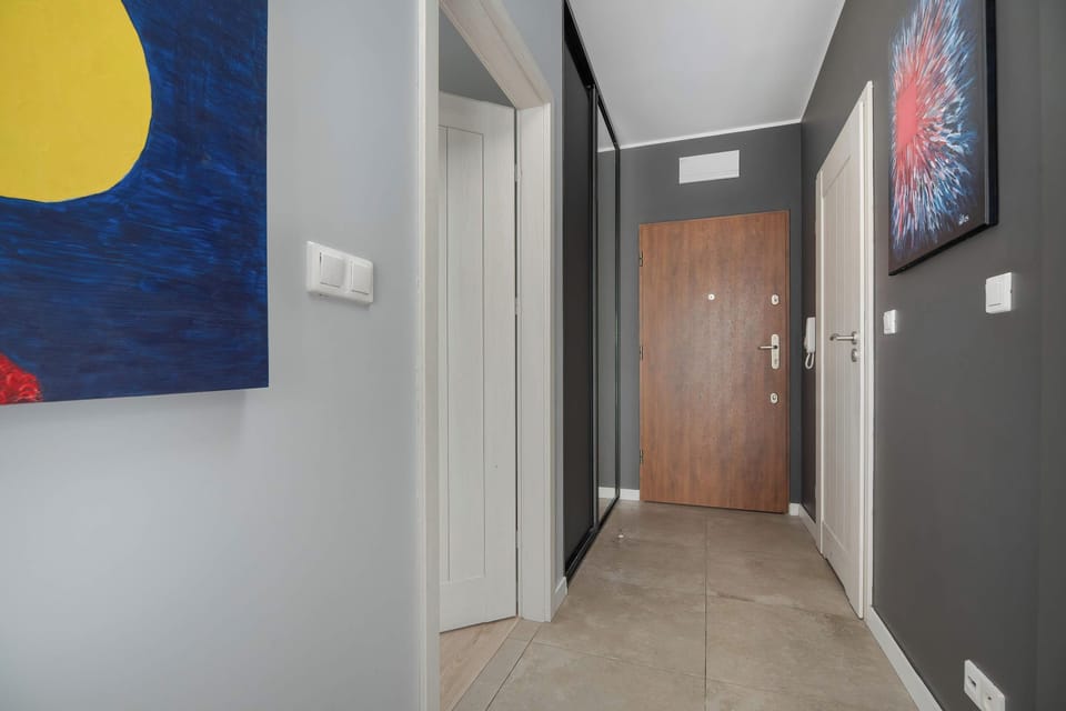A bright apartment hallway with a wooden entrance door, gray walls, and colorful wall art.