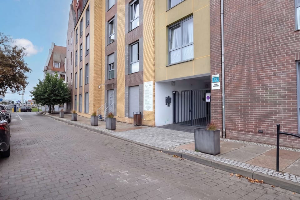 The exterior of the apartment building viewed from a quiet paved street. The modern brick facade and surrounding greenery create a pleasant residential setting.