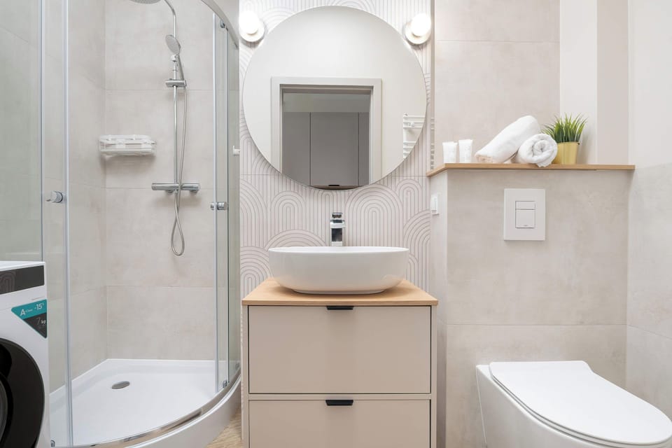 A bright bathroom scene features a glass-enclosed corner shower cabin beside a wooden vanity with a vessel sink. A round mirror with integrated lighting hangs above the sink, reflecting the clean, tiled walls.