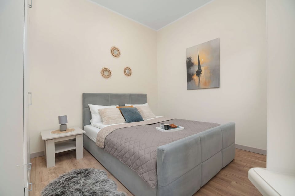 A bright sleeping area offers a double bed with an upholstered headboard, dressed in neutral linens and decorative pillows. A small nightstand with a lamp sits beside the bed, complemented by minimalist wall decor.