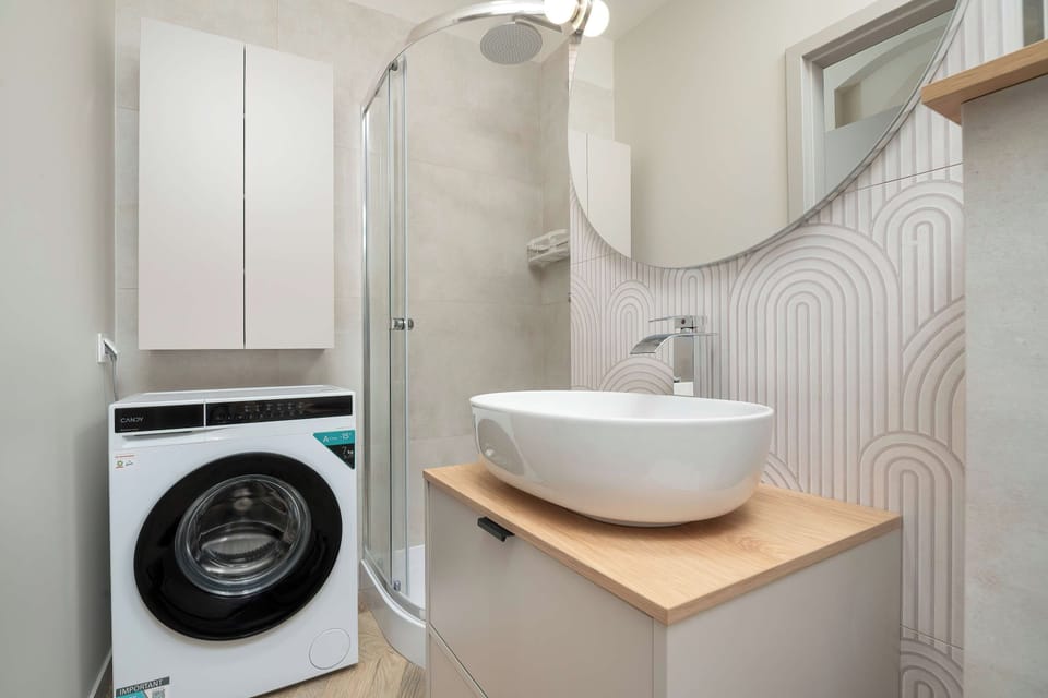 A contemporary bathroom setup presents a front-loading washing machine tucked neatly beneath white utility cabinetry. Next to it, a vessel sink rests on a light wood countertop under an illuminated round mirror.
