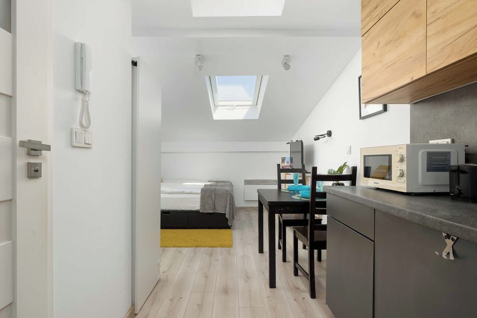 A bright hallway leads into a combined living and sleeping area under a slanted ceiling. A kitchen counter is visible on the right, while a bed and dining table can be seen further into the room.