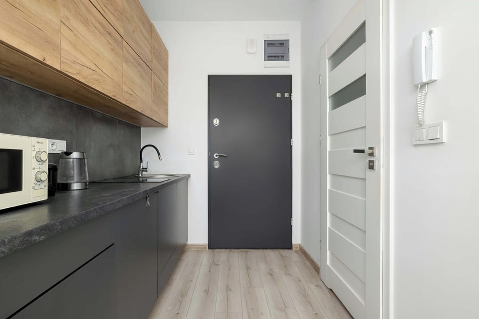 A view down a hallway shows a modern kitchenette with dark lower cabinets and wooden upper shelving. A closed gray door stands at the end of the corridor near a white frame door. The minimalist design utilizes contrasting dark and light elements.
