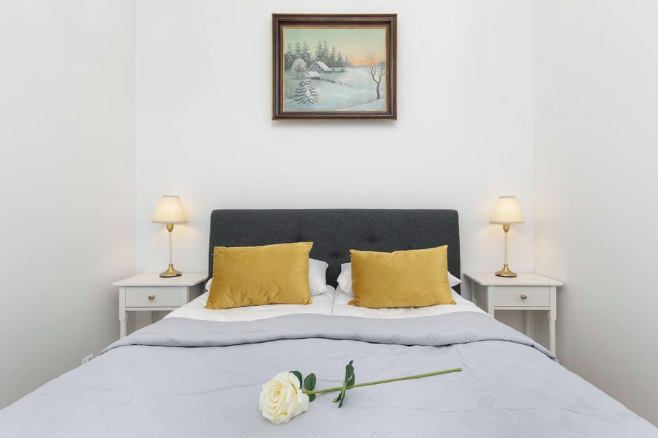 A comfortable bedroom with a double bed dressed in light bedding and accented with yellow pillows. A framed picture and bedside lamps add simple decorative touches.