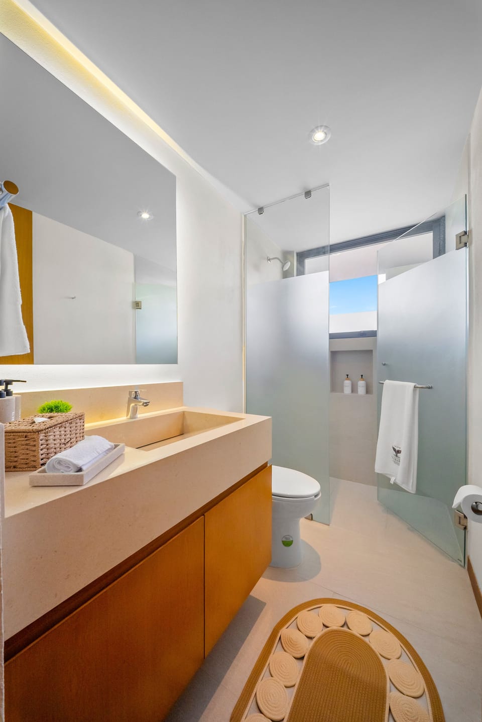 Bright, modern bathroom featuring a stone floating vanity, large backlit mirror, warm wooden cabinetry, glass walk-in shower with frosted panels, and clean coastal finishes.