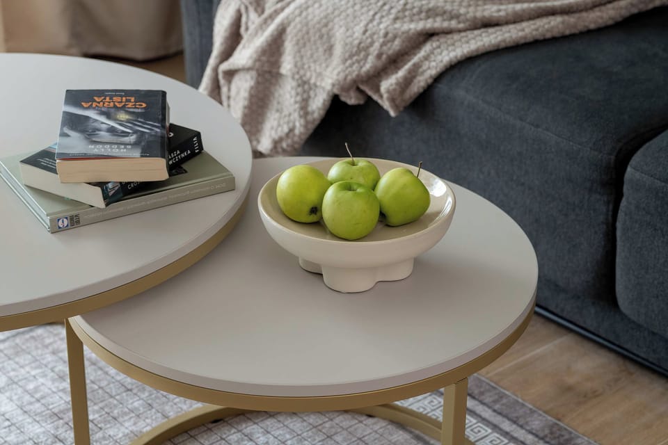 A close-up of a round coffee table with a bowl of green apples and a few decorative items. The table is positioned next to a comfortable dark gray sofa.