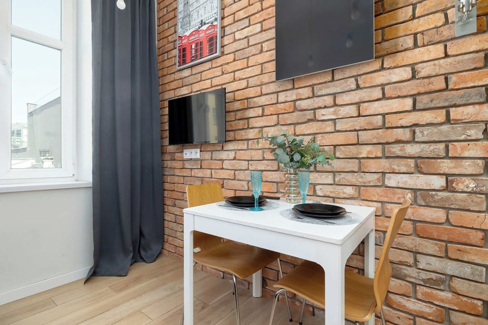 A compact workspace with a wooden desk and chair set against a brick wall. A decorative plant and wall art add character to the bright studio interior.