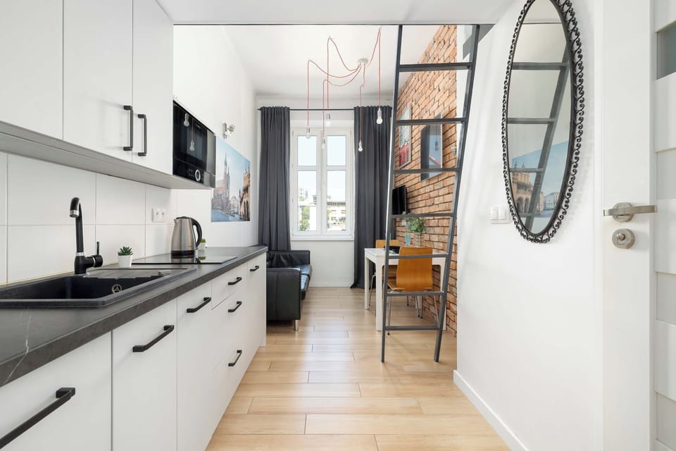 A view of the kitchenette with white cabinetry, a dark countertop, and a sink. A round wall mirror and the studio entrance are visible beside the kitchen area.
