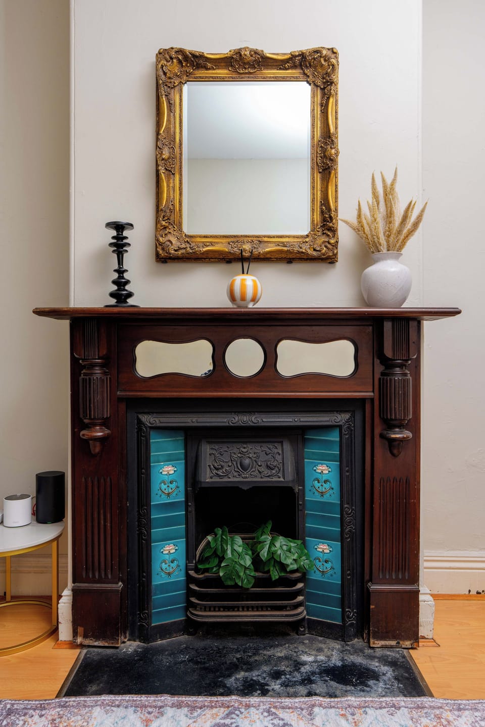 Detail the vintage dark timber fireplace mantel showcasing its decorative blue tile surround, an ornate gilt-framed wall mirror, and dried botanical styling.