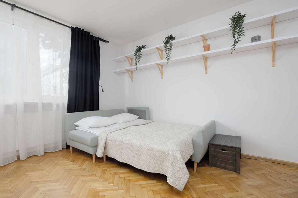 A comfortable sofa bed with neatly arranged bedding is positioned next to large windows. The simple decor and soft colors create a welcoming place to relax and sleep.