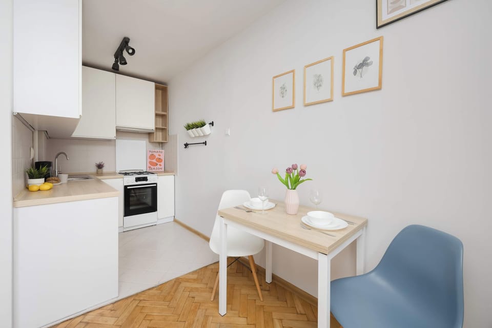 A compact dining area with a small table and two chairs is located next to the kitchenette. The bright space features light furnishings and decorative details on the walls.