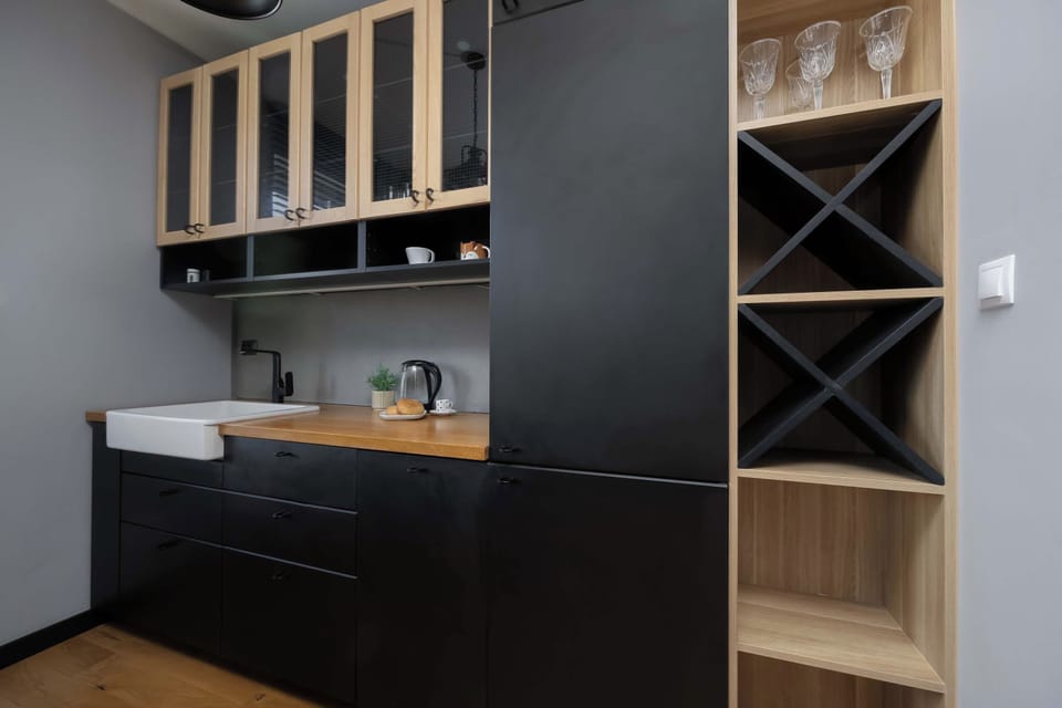 A close-up view of a modern kitchenette with sleek black cabinetry and open wooden shelving. A pristine white sink and contemporary fixtures complete the elegant, minimalist design.
