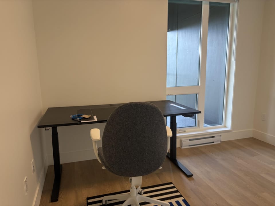 Black adjustable desk with gray swivel chair sits beside tall window with white trim and light wood floors.
