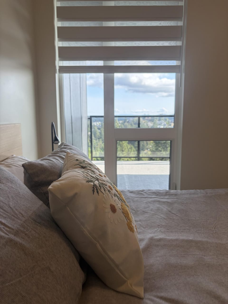 Light filters through layered blinds onto a bed with embroidered floral pillows near glass doors opening to a balcony view.