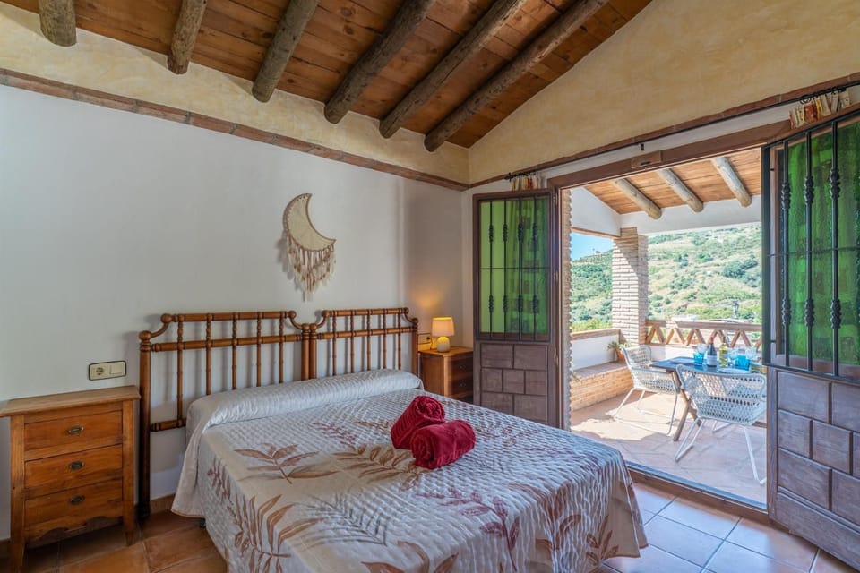 Rustic beach villa bedroom featuring a bamboo headboard, traditional exposed log ceiling beams, and wooden double doors that open directly onto a private dining terrace with scenic hillside views.
