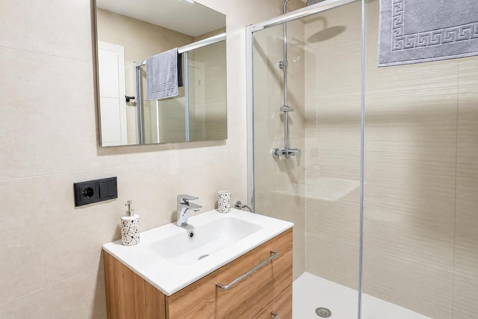 Modern beach villa bathroom featuring a warm wood vanity with a white ceramic sink, a large mirror, and a glass-enclosed walk-in shower with a rain shower head.