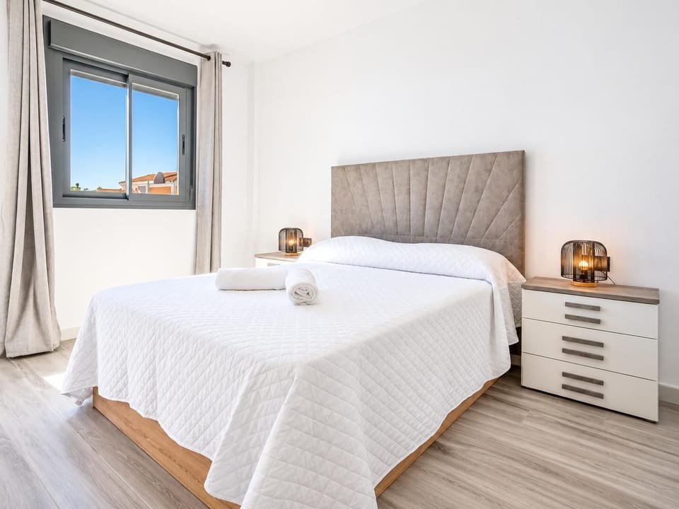 Bright and modern beach villa double bedroom, featuring a minimalist fluted headboard, fresh white linens, contemporary bedside tables, and a large window under a blue sky.