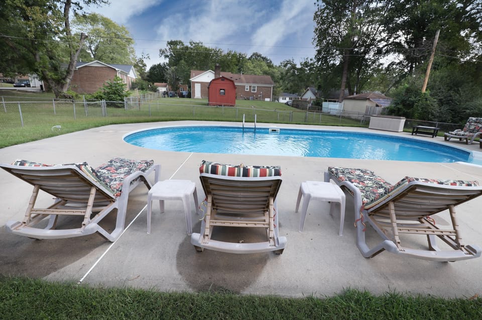 An outdoor swimming pool and patio with cushioned lounge chairs in a fenced yard.