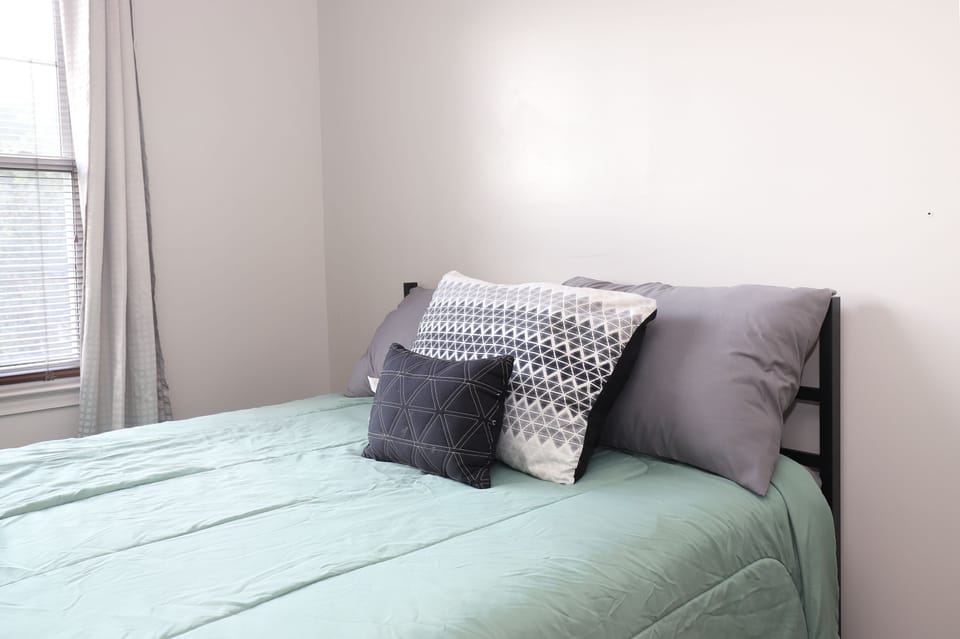 A neatly made bed with geometric throw pillows sits beside a window with blinds.