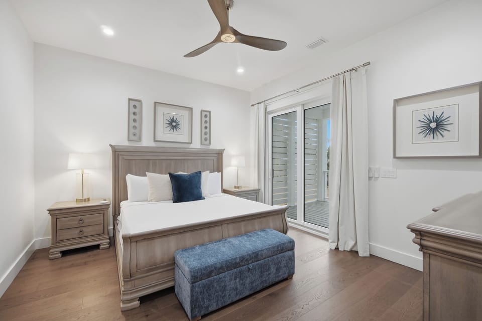 Slip into crisp white linens in your serene bedroom, where soft natural light filters through shuttered doors and a gentle ceiling fan keeps you cool. The elegant coastal touches and plush storage bench create your perfect retreat.
