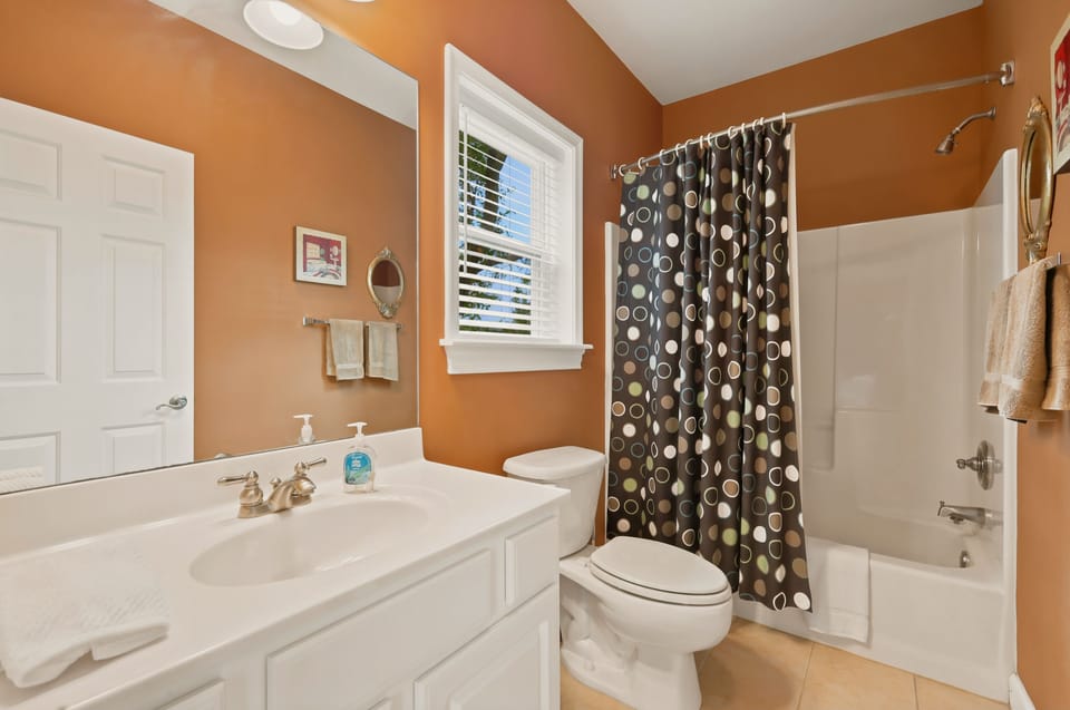 Warm orange walls and a polka-dot shower curtain give this full bath some personality. There is a tub and shower combination, a white vanity under a big mirror, and a window with blinds for daylight, with fresh towels hung and ready to use.