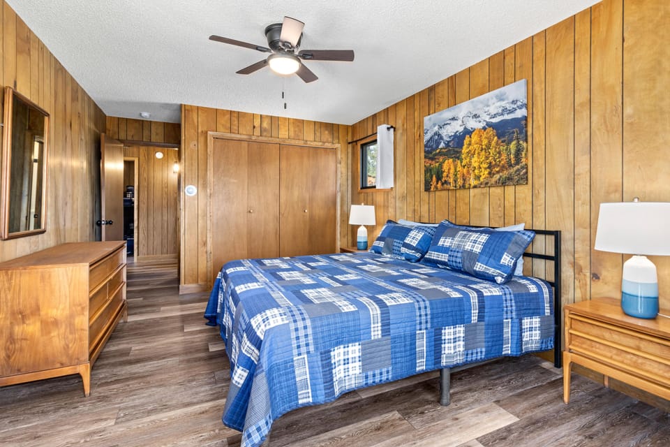 Wood-paneled bedroom with blue plaid bedding, mid-century modern wood dresser, and mountain landscape artwork.