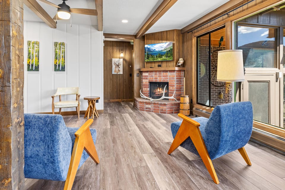 Blue mid-century chairs with wood arms face a brick fireplace under a wall-mounted TV beside large windows and a floor lamp.