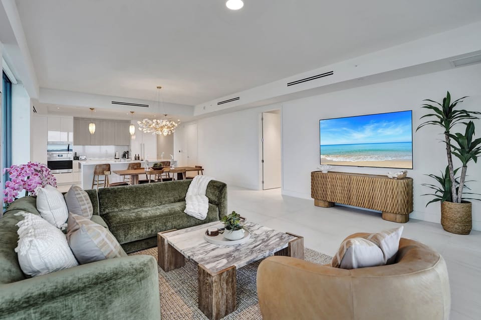 Spacious living room with velvet sofa, marble coffee table, large TV, and open kitchen visible behind.