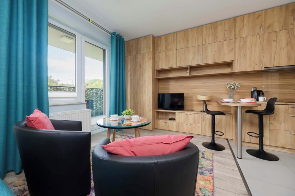 A comfortable studio interior with a double bed, kitchenette, and two armchairs. The wooden cabinetry and blue curtains create a warm and inviting atmosphere.