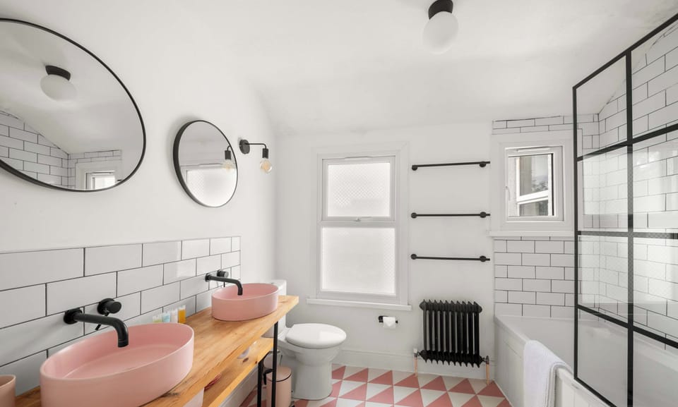 A modern bathroom features a wooden vanity with two pink vessel sinks, round black-framed mirrors, a white bathtub with a black grid shower screen, and a geometric pink-and-white tiled floor.