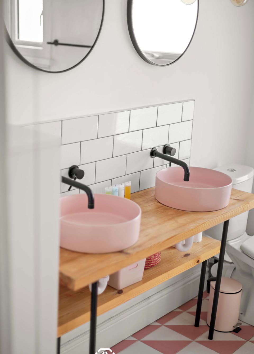 A modern bathroom features two pink vessel sinks with black wall-mounted faucets on a wooden vanity, complemented by round mirrors, white subway tiles, and a pink patterned floor.