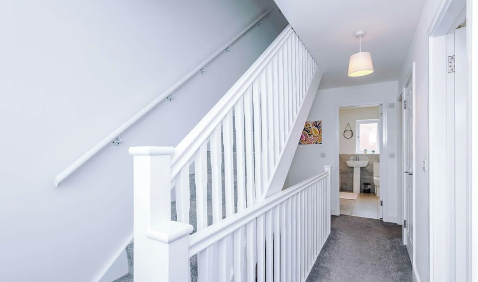 This bright hallway features a white wooden staircase with a matching banister, grey carpeting, a hanging light fixture, and an open doorway down the hall that reveals a partial view of a modern bathroom.