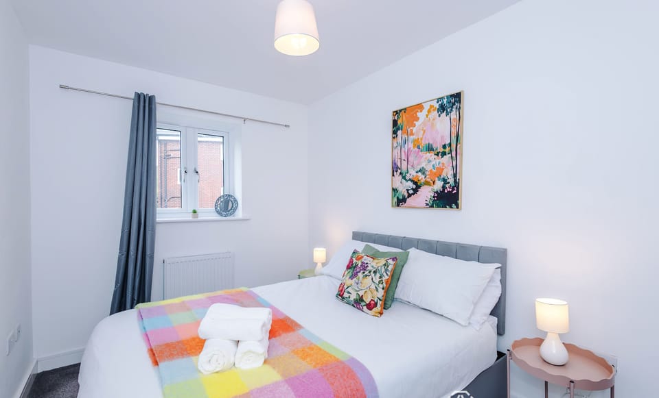 This bright bedroom features a neatly made double bed dressed with a vibrant multicolored plaid throw and rolled white towels, alongside a small window with grey curtains, colorful abstract wall art, and twin glowing bedside lamps.