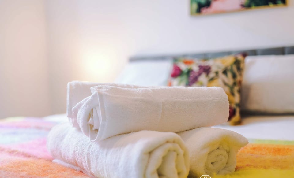 A neat stack of rolled white bath towels resting on a colorful blanket, with a softly blurred bed, a floral accent pillow, and warm background lighting.