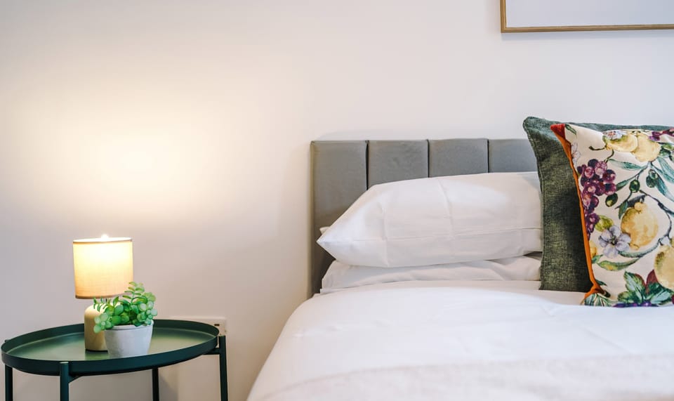A cozy bedroom setup highlights a bed with white linens, decorative patterned pillows, and a grey tufted headboard, next to a small round bedside table with a glowing lamp.