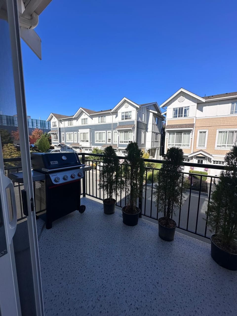 Balcony with speckled flooring features a black gas grill and four potted evergreen shrubs.