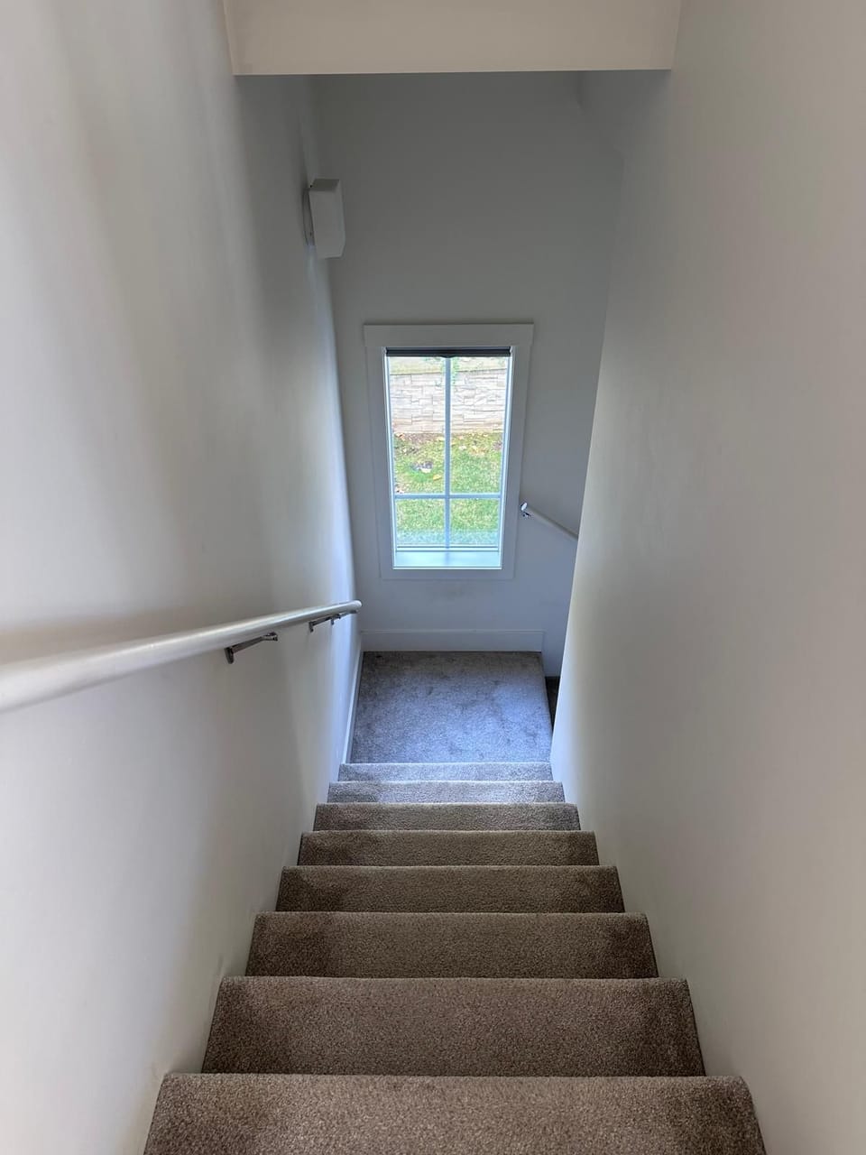 Carpeted staircase with white walls leads down to a window overlooking a stone retaining wall and grass.