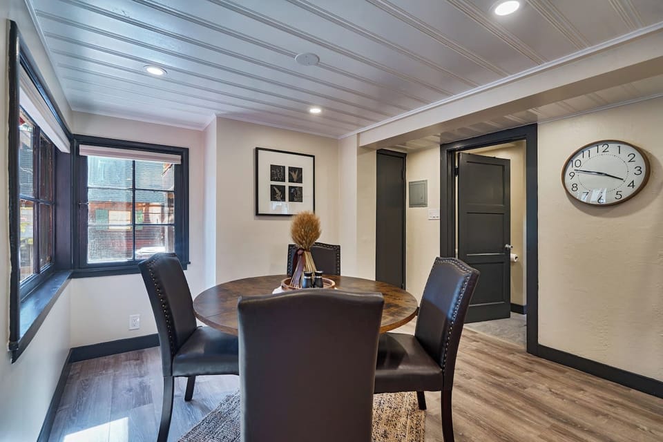 A round wood table sits framed by black-trimmed windows, with charcoal doors and warm hallway lighting just beyond. Simple and inviting, it`s a versatile dining spot bathed in natural light.