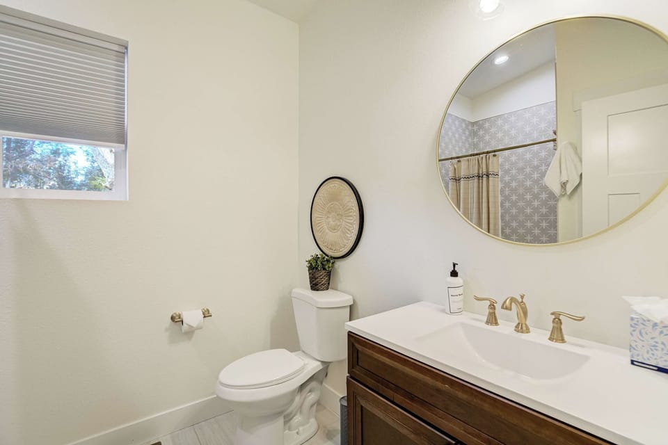 Brushed bronze fixtures warm the primary en suite, set against a dark wood vanity and white counter beneath a round gold framed mirror. A small shaded window lets in soft light, and the walk-in shower waits behind a curtain at the far end.