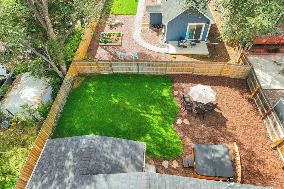 From above, the lawn reads as one green rectangle bordered by mulch beds, with the patio, dining set, and hot tub tucked along the near side of the house. Trees crowd the far edge, and the parking pad sits just beyond the other end of the home.